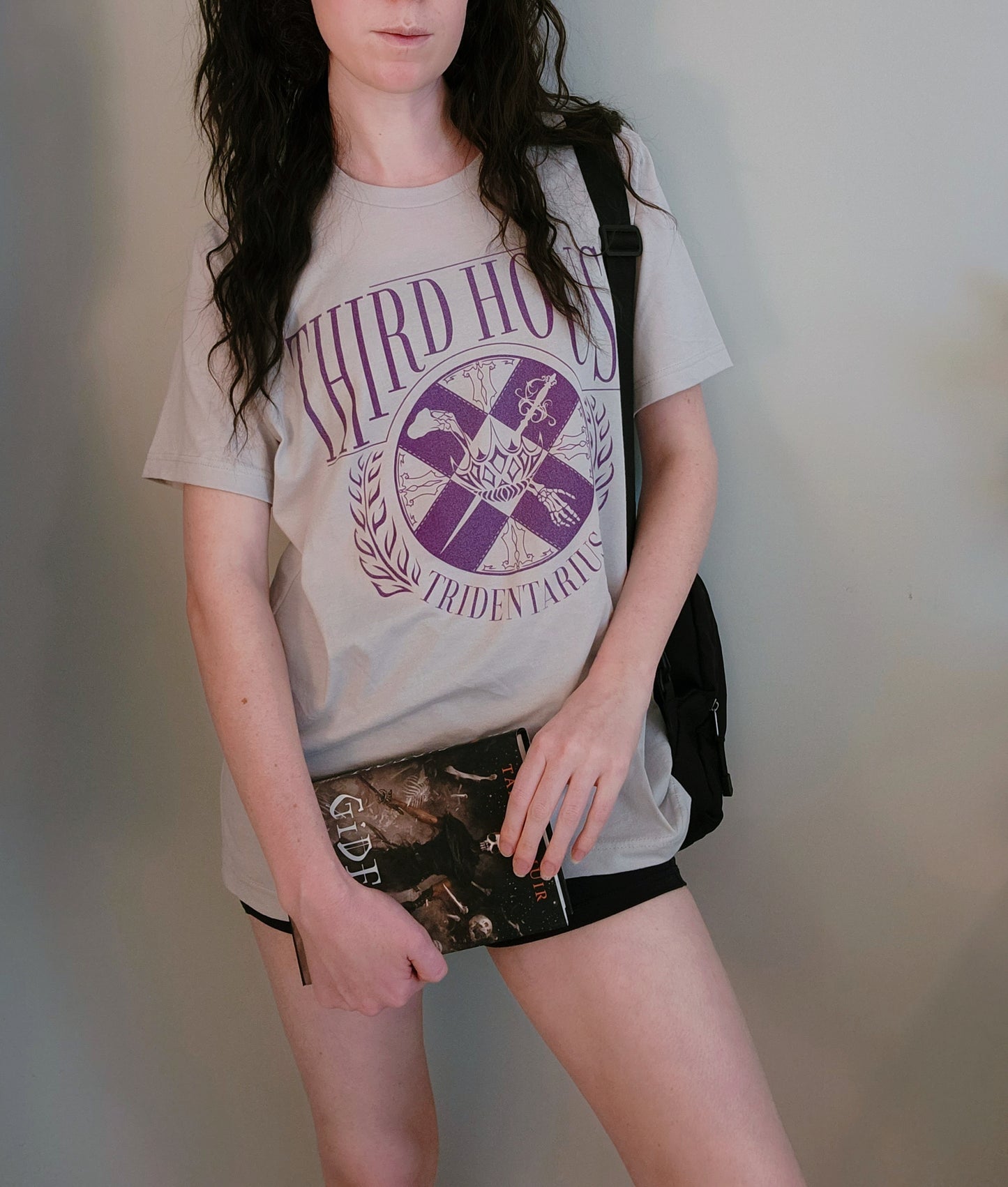 The locked tomb gideon the ninth third house shirt in grey on a model holding a copy of harrow the ninth