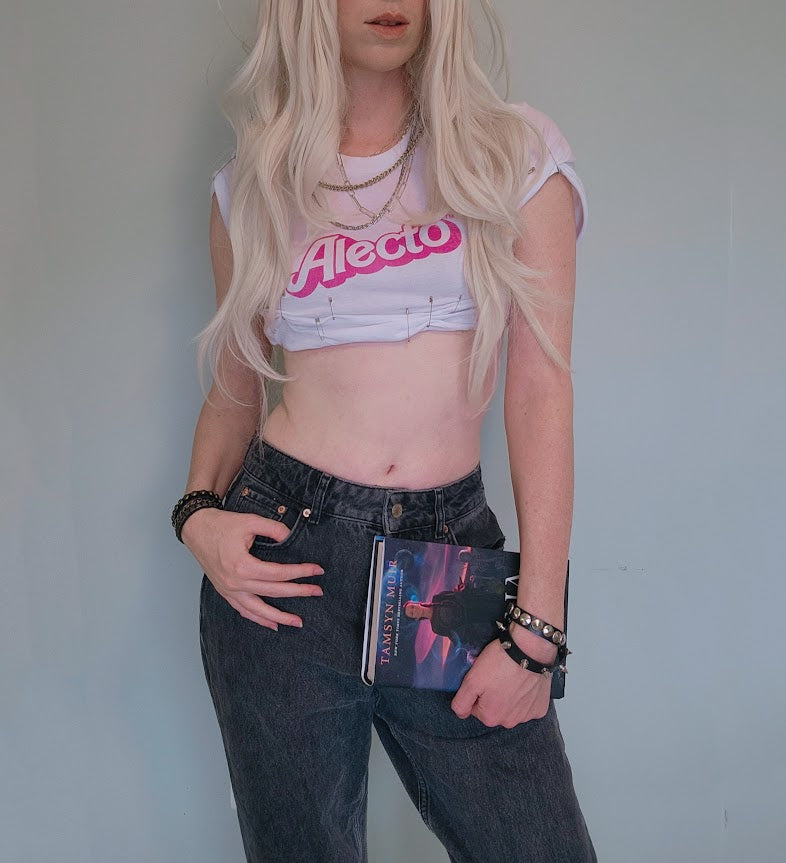 Person wearing a white crop top with pink text the says "Alecto" in the Barbie font and dark jeans, holding a copy of the book nona the ninth from the locked tomb series against a plain background