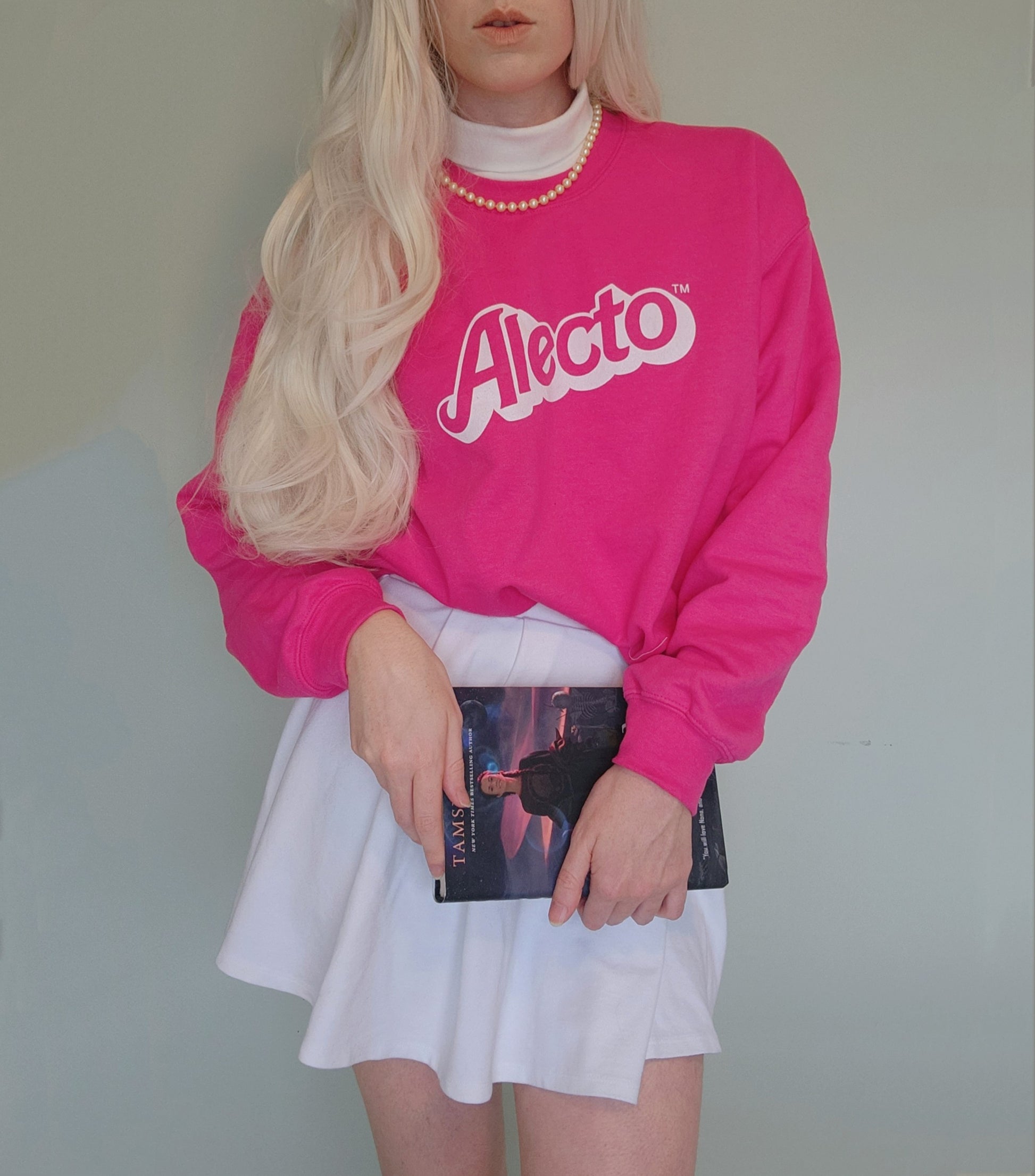 Person wearing a bright pink sweatshirt with 'Alecto' logo, holding a copy of the book nona the ninth, against a plain background