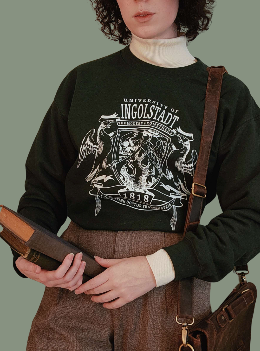 Person wearing a black sweater with a Frankenstein design, holding a brown bag and a copy of Mary Shelley's Frankenstein against a plain background