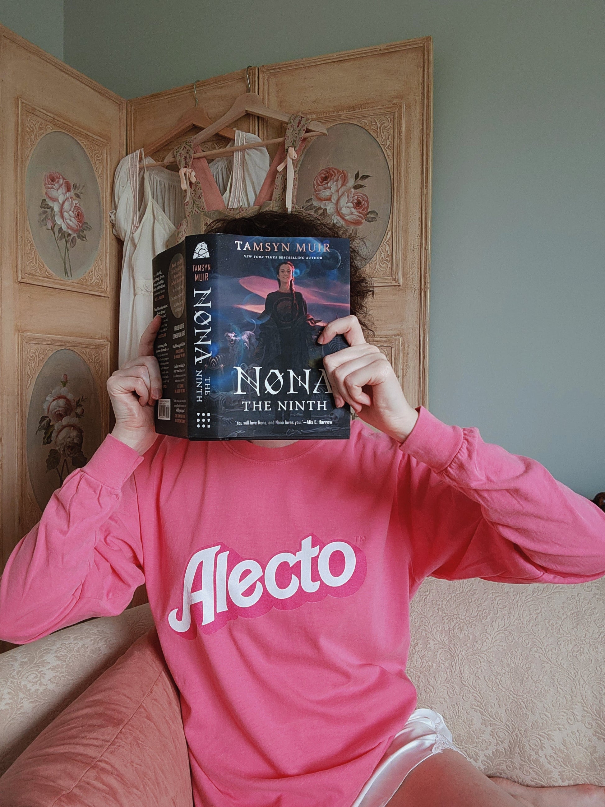 Person wearing a pink long sleeve shirt with 'Aleco' text, holding a copy of the book Nona the Ninth in front of their face.